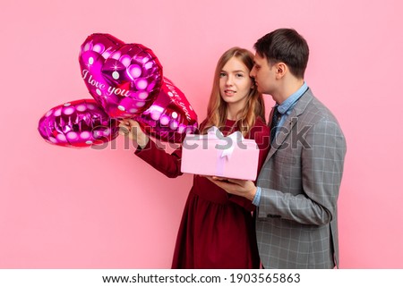 Beautiful loving young couple cuddling having fun together celebrating Valentine's Day with heart shaped balloons and gift box on pink background