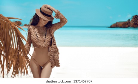 Beautiful luxury portrait of brunette woman in beige hat, boho swimsuit posing by ocean water. Sexy tanned body, perfect figure. Girl model resting on tropical island with copyspace background.