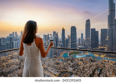 A beautiful luxury woman in a white dress enjoys the sunset view behind the illuminated skyline of Downtown Dubai, UAE, with a drink - Powered by Shutterstock