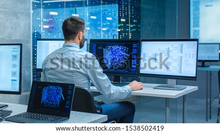 Beautiful Male Computer Engineer and Scientists Create Neural Network at His Workstation. Office is Full of Displays Showing 3D Representations of Neural Networks.