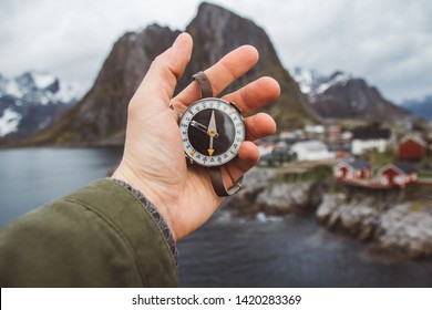 Beautiful male hand holds a magnetic compass against the background of the houses and mountains rocks over fjord landscape . The concept of finding yourself the way and the truth. - Shutterstock ID 1420283369