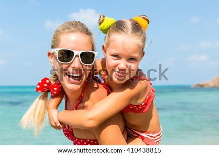 Beautiful mammy sunglasses with cute little daughter on her back. Two in red bikini. Woman and little lady smile and looking to camera. Happy family on the sea shore. Happy mothers day.
