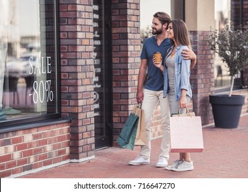 Beautiful man and woman are carrying shopping bags, looking at shop window and smiling while walking down the street