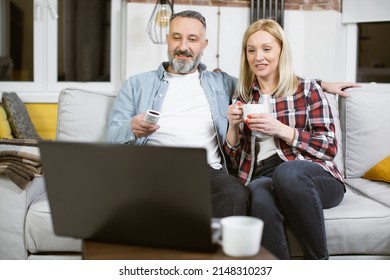 Beautiful Married Couple Of Two Mature People In Casual Wear Sitting Together On Couch And Watching Film On Modern Laptop. Happy Caucasian Family Spending Free Time Together At Home.