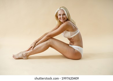 Beautiful Mature Blonde Woman With Fit Body Posing In Underwear, Leaning On Knees While Sitting On Studio Floor And Smiling At Camera Isolated Over Beige Background