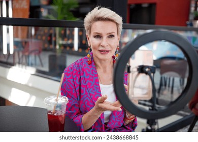 Beautiful mature blonde woman with a short haircut, an influencer, records a video blog while sitting at a table in a restaurant. Middle aged female blogger. - Powered by Shutterstock