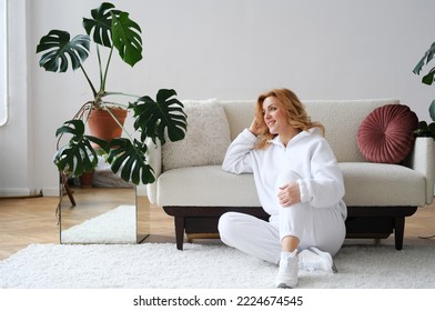 A beautiful mature blonde woman sits in a living room on a carpet in a tracksuit. Woman smiling and looking out the window. Horizontal photo