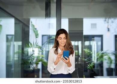 Beautiful mature business woman executive using smartphone walking in office. Smiling elegant mid aged businesswoman manager holding mobile cell phone looking at cellphone tech standing in lobby. - Powered by Shutterstock