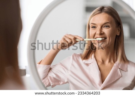 Beautiful Mature Female Brushing Her Teeth With Toothbrush Near Mirror, Happy Middle Aged Woman Smiling At Her Reflection, Making Morning Hygiene At Home, Doing Oral Care Treatments, Selective Focus