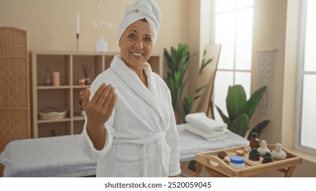 A beautiful mature hispanic woman in a spa room wearing a bathrobe and towel turban, smiling and extending her hand in a welcoming gesture. - Powered by Shutterstock