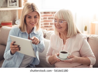 Beautiful mature mother and her adult daughter are drinking coffee, using a digital tablet and smiling while sitting on couch at home - Powered by Shutterstock