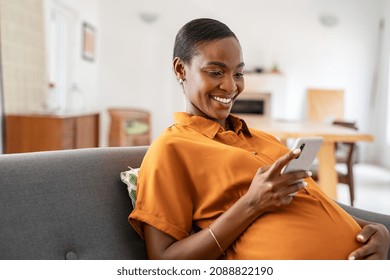 Beautiful mature pregnant woman watching funny video on smartphone. Happy black pregnant woman using mobile while touching tummy at home. Middle aged expecting african american woman using cellphone.
