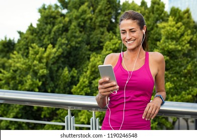 Beautiful mature runner chatting on mobile phone while relaxing after fitness training outside. Latin woman using smartphone screen while listening to music on earphones after jogging in the city. - Powered by Shutterstock