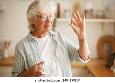 Beautiful Mature Sixty Year Old Female In Stylish Eyeglasses Holding Mug And Omega 3 Supplement Capsule, Going To Take Vitamin After Meal. Senior Gray Haired Woman Taking Fish Oil Pill With Water