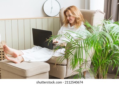 Beautiful Mature Smiling Woman Working On Laptop And  Sitting In A Big Comfortable Chair At Home. Female Entrepreuneur And Freelance Concept