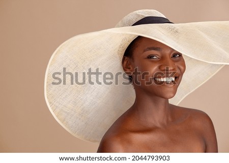 Beautiful mature woman with bare shoulder wearing summer hat with large brim and looking at camera. Portrait of smiling attractive black woman wearing straw hat with a wide brim isolated on background