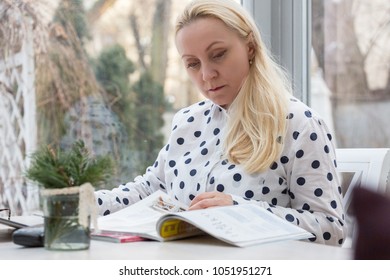 Beautiful mature woman - business woman with blond hair loose, works sitting in a coffee shop by reading a magazine. Bright interior.