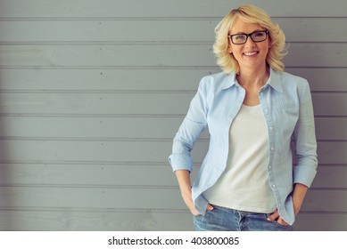 Beautiful Mature Woman In Casual Clothes And Eyeglasses Is Looking At Camera And Smiling, Standing Against Gray Background