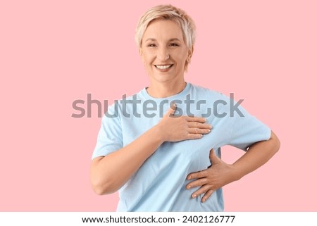 Beautiful mature woman checking her breast on pink background. Cancer awareness concept