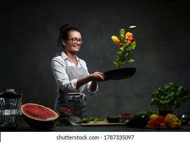 Beautiful mature woman cooking vegetable salad in the kitchen, tosses vegetables from the pan. Healthy food concept. Studio photo on a dark background