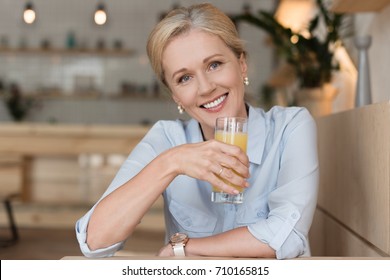 Beautiful Mature Woman Drinking Orange Juice And Smiling At Camera In Cafe