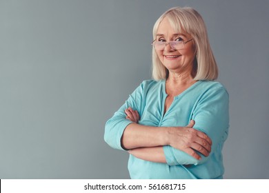 Beautiful Mature Woman In Eyeglasses Is Looking At Camera And Smiling While Standing With Folded  Arms On Gray Background