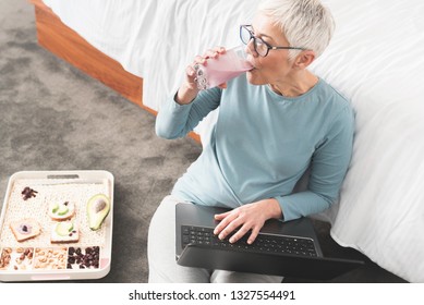 Beautiful Mature Woman With Glasses Drinking Fruit Smoothie For Energy And Searching Online On Her Laptop, Relaxing And Enjoying Free Time Concept