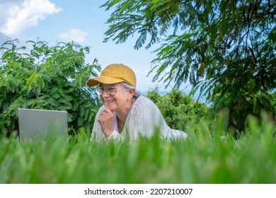 Beautiful Mature Woman With Hat And Eyeglasses Lying Down In The Meadow Looking At Laptop Smiling. Elderly Lady Relaxed In Public Park Enjoying Free Time Or Retirement