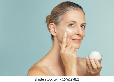 Beautiful Mature Woman Holding Jar Of Skin Cream For Face Isolated On Light Blue Background. Happy Senior Woman Applying Anti-aging Moisturizer And Looking At Camera. Beauty And Anti Aging Treatment.