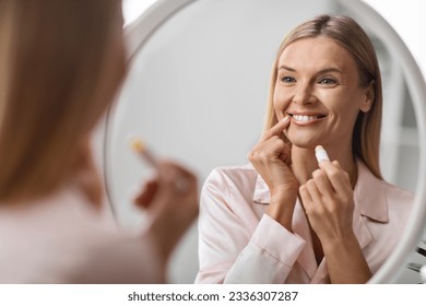 Beautiful Mature Woman Looking At Mirror And Applying Moisturising Lip Balm, Smiling Attractive Middle Aged Lady Holding Chapstick, Using Hygienical Lipstick While Making Domestic Beauty Routine - Powered by Shutterstock