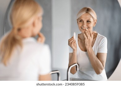 Beautiful Mature Woman Looking At Mirror And Applying Moisturising Lip Balm, Attractive Happy Senior Lady Holding Chapstick, Using Hygienical Lipstick While Making Domestic Beauty Routine - Powered by Shutterstock