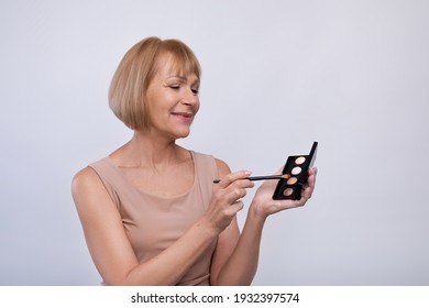 Beautiful Mature Woman With Silky Skin Applying Eye Shadow, Holding Palette On Light Studio Background, Copy Space. Senior Lady Using Natural Cosmetic Product, Taking Care Of Her Looks