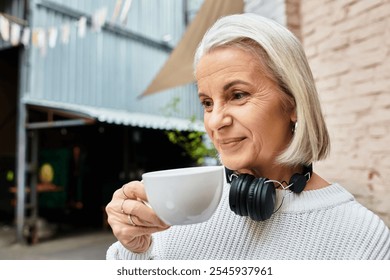 A beautiful mature woman sips coffee and smiles serenely while wearing headphones outside. - Powered by Shutterstock