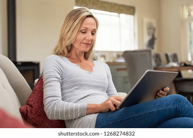 Beautiful mature woman sitting on couch and using digital tablet. Smiling lady browsing internet over tablet at home. Middle age woman holding computer at home.