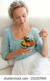 Beautiful Mature Woman Sitting And Relaxing On A White Sofa At Home While Eating A Small Green Salad, Home Interior. Senior Woman Eating Healthy Food, Wellness And Well Being Indoors. Lifestyle.