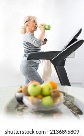 Beautiful Mature Woman In Sportswear Drinking Water From Thermos Bottle While Jogging On The Treadmill At Home. Seniors, Physical Activity, Home Workout And Health Lifestyle Concept.