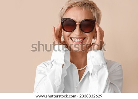 Beautiful mature woman in sunglasses on beige background, closeup