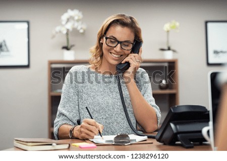 Beautiful mature woman talking on phone at creative office. Happy smiling businesswoman answering telephone at office desk. Casual business woman sitting at desk making telephone call and taking note.