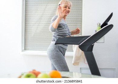 Beautiful Mature Woman Waving Hand To The Camera While Jogging On Treadmill At Home.