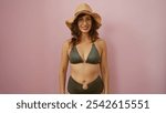 Beautiful mature woman wearing a bikini and a straw hat smiling confidently against a pink isolated background