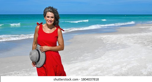 Beautiful Mature Woman Wearing A Red Summer Dress And Holding Her Hat On The Beach