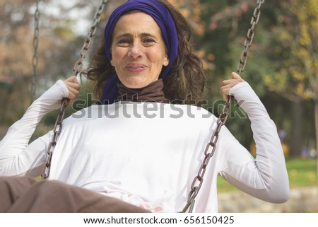 Beautiful mature woman with white long sleeve shirt and purple shawl on head swinging in the playground. Middle aged pretty lady at park. Child memories, young at heart concepts