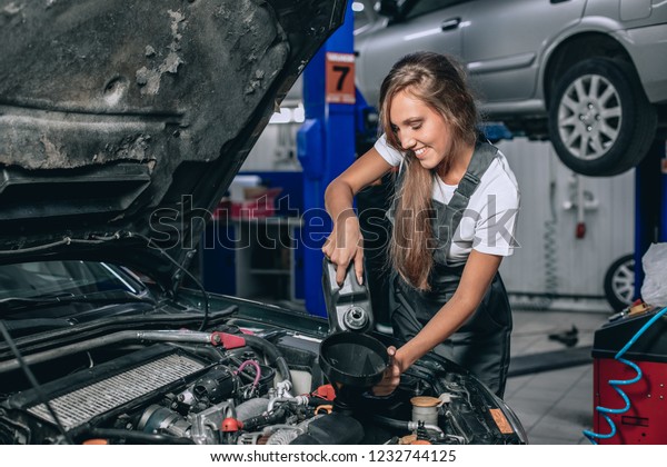 Beautiful Mechanic  girl in a black jumpsuit and\
a white T-shirt changes the oil in a black car and smiling at the\
camera. car repair\
concept
