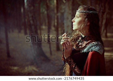 A beautiful medieval queen stands in a dense forest looking at the light breaking through the branches of the trees. Ancient legends. Fantasy world.