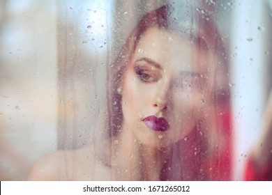 A beautiful melancholic alone sad woman stands in front of the window, looking down thinking in sadness thoughtful. Image made through the window, rain on window and reflection on outdoors scene. 