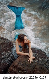 Beautiful mermaid girl on the seashore. Mermaid on the ocean against the background of rocks. Mermaid with a blue tail on the beach. Mermaid on the background of the waves. Siren washed ashore.