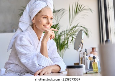 Beautiful mid age adult 50 years old blonde woman wears bathrobe and towel in bathroom applying rejuvenating tightening antiage face skin care cream treatment, sitting at mirror. Daily beauty routine.