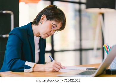 Beautiful Middle Age Asian Woman Working On Paperwork, Sitting In Modern Contemporary Office, With Laptop Computer. Business Owner, Entrepreneur, Executive Manager, Or Employee Office Worker Concept