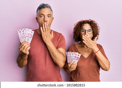 Beautiful Middle Age Couple Holding 50 Mexican Pesos Banknotes Covering Mouth With Hand, Shocked And Afraid For Mistake. Surprised Expression 