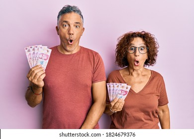 Beautiful Middle Age Couple Holding 50 Mexican Pesos Banknotes Scared And Amazed With Open Mouth For Surprise, Disbelief Face 